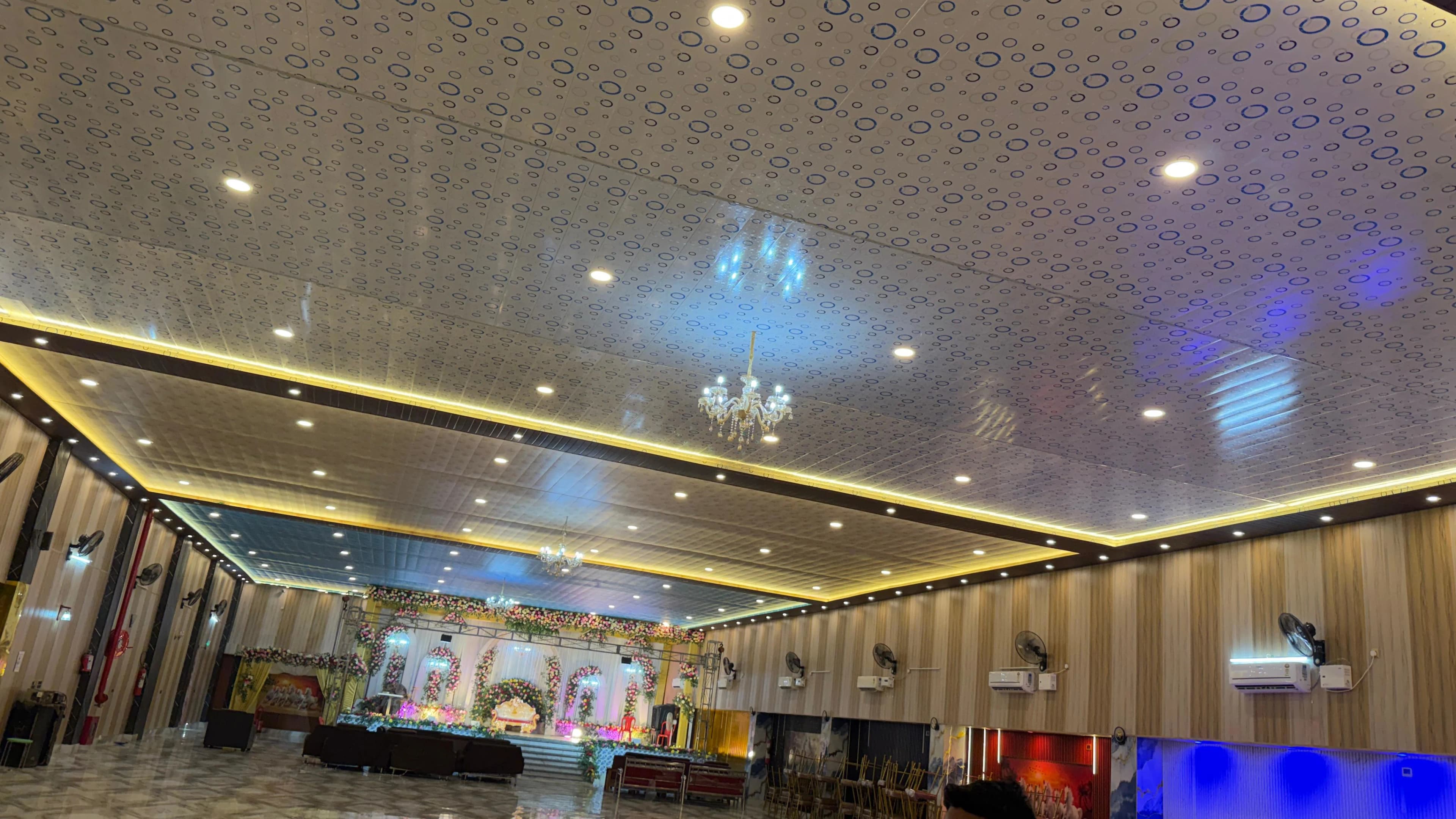 Wide view down the hall towards the flower-decked stage, chandeliers and lit cove ceiling overhead and banquet chairs along one wall