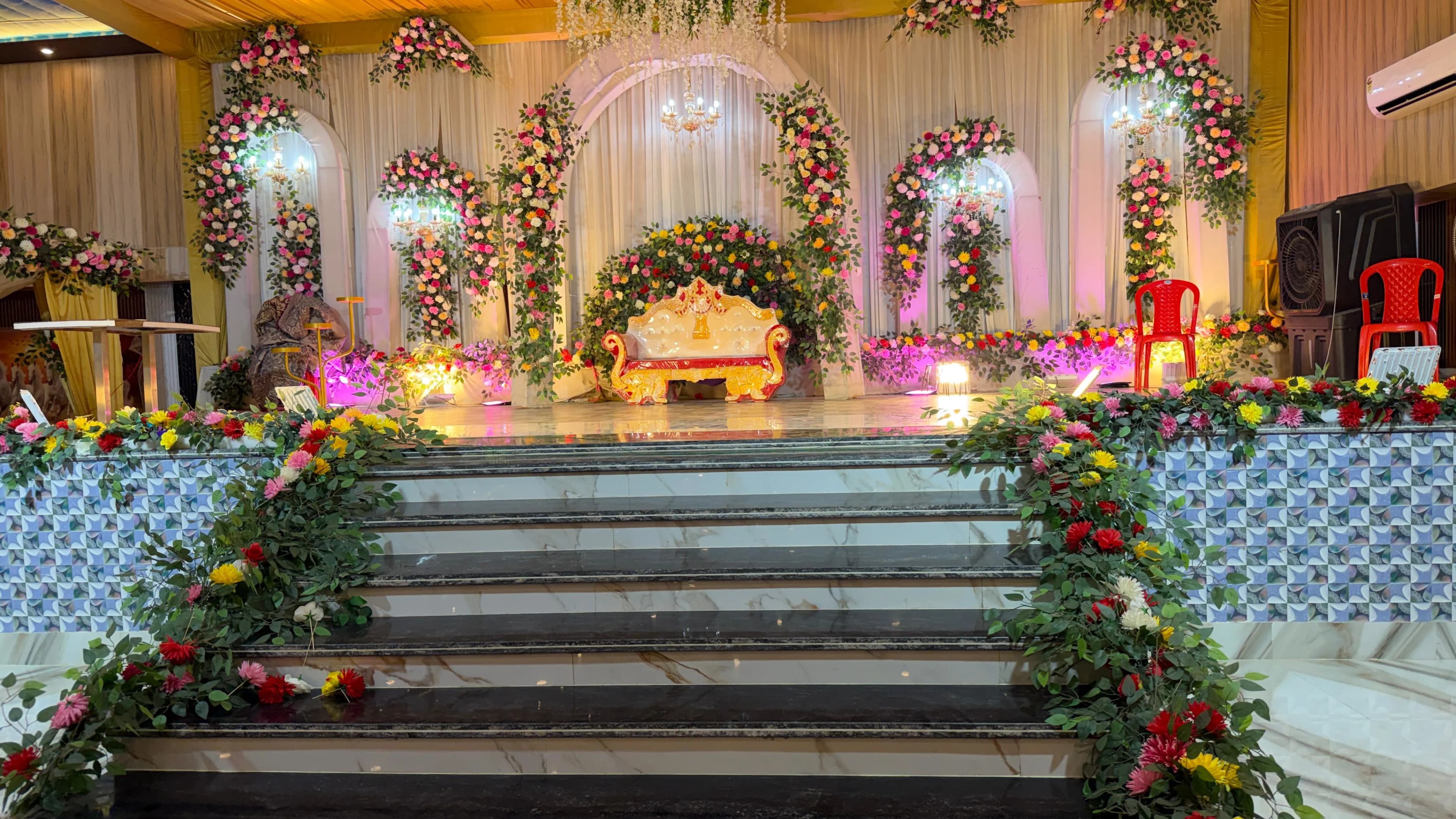 The reception stage head-on — a gilded settee beneath floral arches and chandeliers, with garlanded marble steps leading up to it